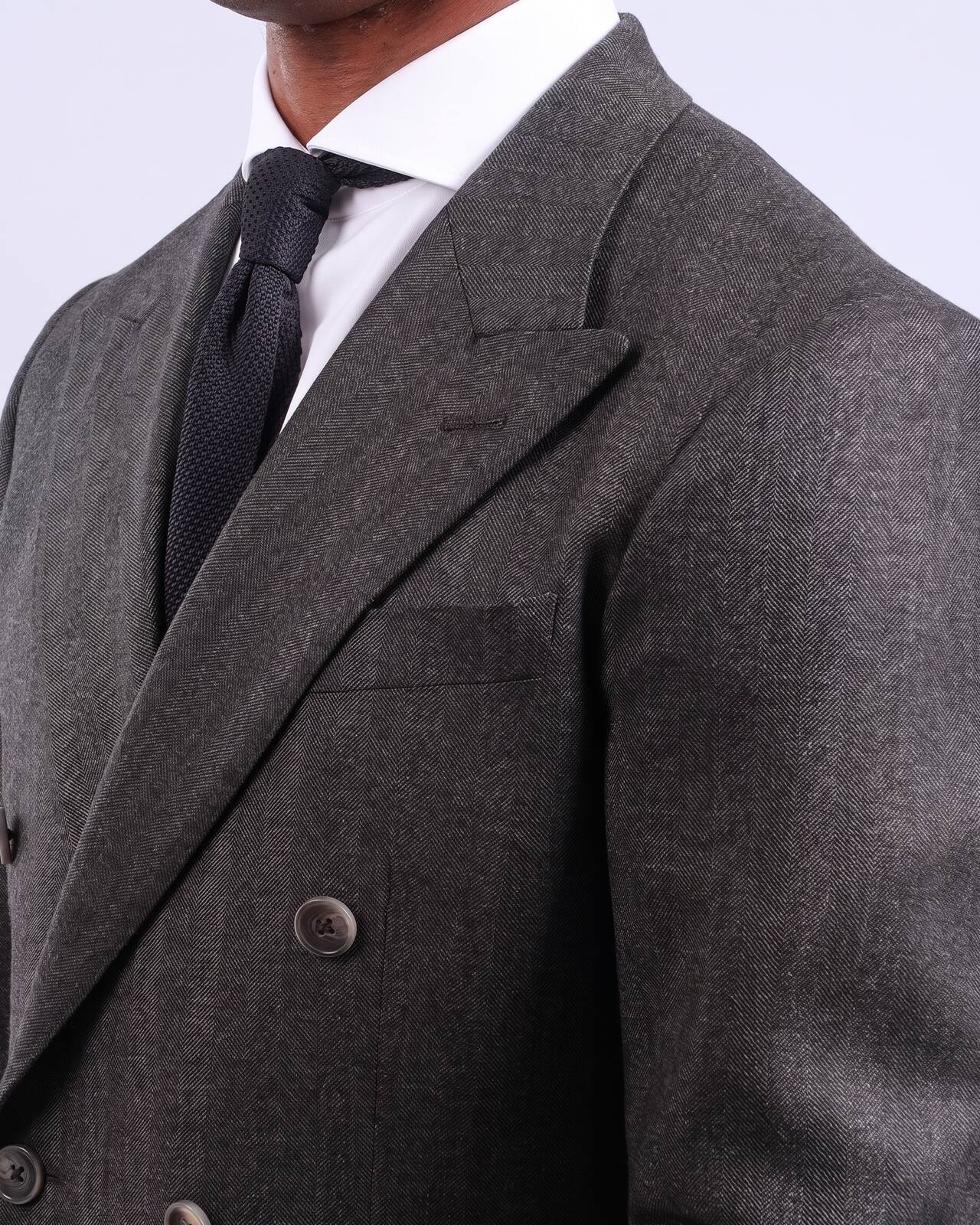 Grey herringbone double-breasted jacket seen close up — peak lapel, welted breast pocket, worn over a white shirt and a navy grenadine tie